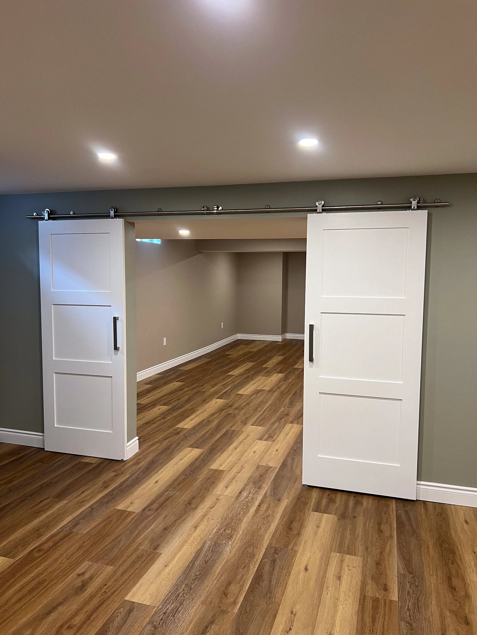 Finished basement with two white barn doors on a steel track, sage accent wall, wood-look plank flooring and recessed pot lights